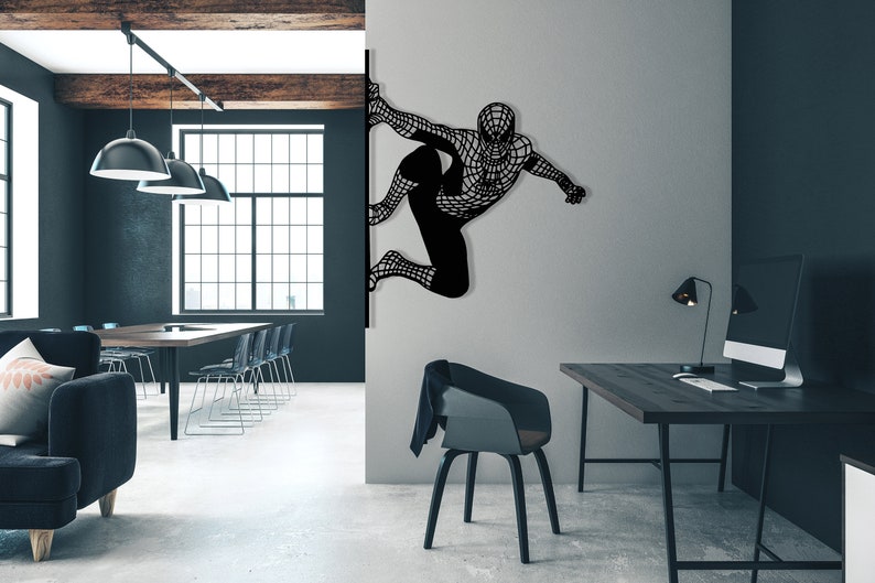 Op de afbeelding: Zwarte metalen wandkunst met een superheld in een web-pak, hangend aan een witte muur in een moderne kantooromgeving. De kunst is gemonteerd op een muur naast een bureau met een computer en een lamp.