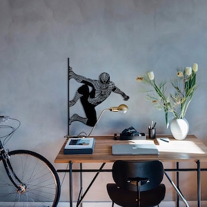 Op de afbeelding: Zwarte metalen wandkunst met een gestileerd silhouet van Spiderman die een muur beklimt. De kunst is gemonteerd op een grijze muur met een bureau en een stoel op de achtergrond.