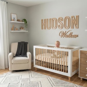 Puede incluir: Un cuarto de beb&eacute; con una cuna blanca, un sill&oacute;n beige y una c&oacute;moda de madera. La pared presenta un letrero de madera que dice "HUDSON Walker". Un beb&eacute; est&aacute; en la cuna. Estantes decorativos con juguetes y plantas.