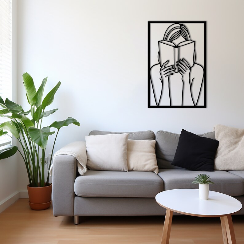 Puede incluir: Una escultura de pared de metal negro de una persona leyendo un libro. La escultura est&aacute; enmarcada en un marco cuadrado negro y est&aacute; colgada en una pared blanca sobre un sof&aacute; gris.