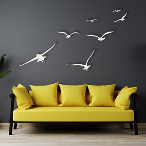 Puede incluir: Un sof&aacute; amarillo con tres almohadas se encuentra frente a una pared gris oscuro con recortes de gaviotas de papel blanco. Una planta en maceta est&aacute; a la izquierda del sof&aacute; y una l&aacute;mpara de pie negra con tr&iacute;pode est&aacute; a la derecha.