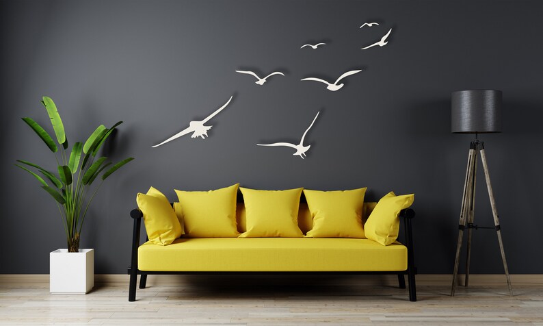 Puede incluir: Un sof&aacute; amarillo con tres almohadas se encuentra frente a una pared gris oscuro con recortes de gaviotas de papel blanco. Una planta en maceta est&aacute; a la izquierda del sof&aacute; y una l&aacute;mpara de pie negra con tr&iacute;pode est&aacute; a la derecha.