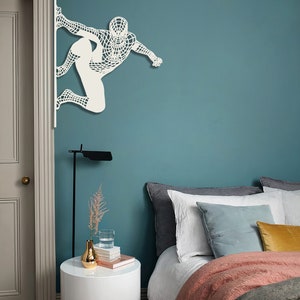 Op de afbeelding: Een tealblauwe muur met een witte uitsnede van Spiderman. De muur is tealblauw. Er is een bed met een witte dekbedovertrek en een roze deken. Er is een wit nachtkastje met een gouden vaas en een stapel boeken.
