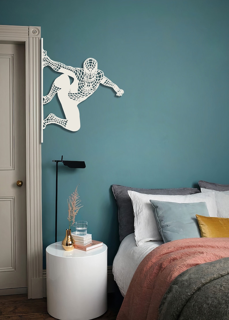 Op de afbeelding: Een tealblauwe muur met een witte uitsnede van Spiderman. De muur is tealblauw. Er is een bed met een witte dekbedovertrek en een roze deken. Er is een wit nachtkastje met een gouden vaas en een stapel boeken.