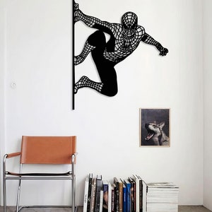 Op de afbeelding: Zwarte metalen wandsculptuur van Spiderman in een hurkende houding, met zijn armen uitgestrekt, alsof hij gaat springen. De sculptuur is gemonteerd op een witte muur, met een bruine leren stoel en een boekenkast op de achtergrond.