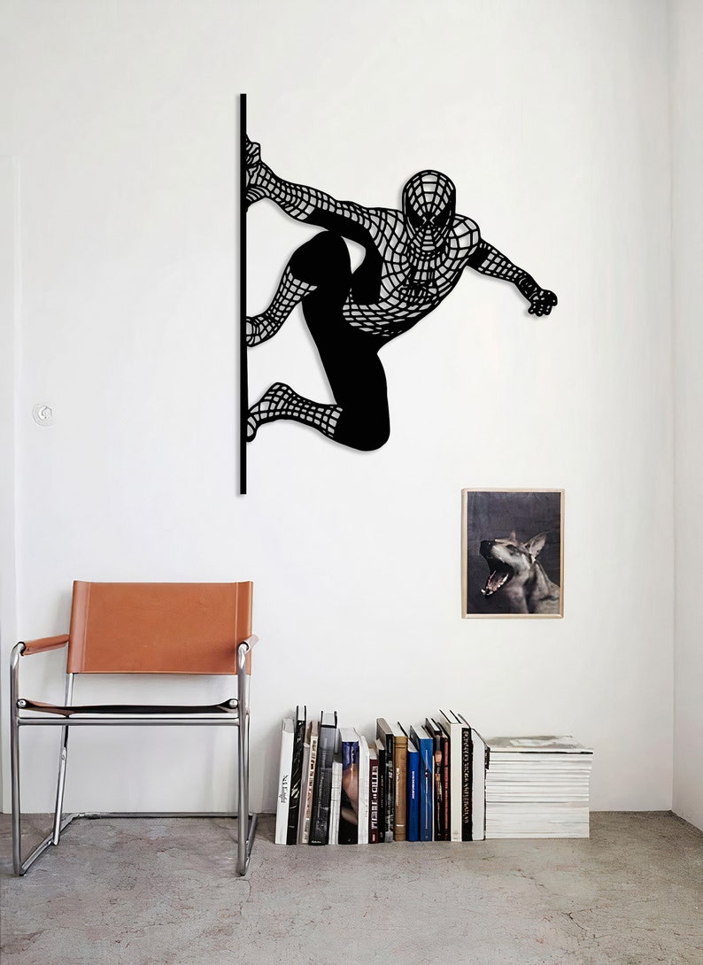Op de afbeelding: Zwarte metalen wandsculptuur van Spiderman in een hurkende houding, met zijn armen uitgestrekt, alsof hij gaat springen. De sculptuur is gemonteerd op een witte muur, met een bruine leren stoel en een boekenkast op de achtergrond.