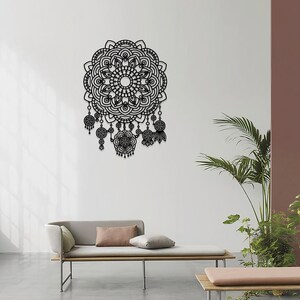 May include: Black metal wall decor featuring a large mandala design with intricate details and dangling charms. The mandala is surrounded by a white wall and a light grey sofa with cushions.