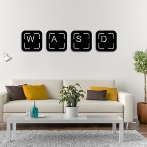 May include: Four black square wall decals with white letters spelling out "WASD" on a white wall. The decals are in a living room setting with a white sofa, a coffee table, and a potted plant.