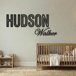 Puede incluir: Decoraci&oacute;n de pared de madera negra con las palabras "HUDSON" y "Walker" en un cuarto de ni&ntilde;os. Tambi&eacute;n se ve una cuna de madera con un oso de peluche y una peque&ntilde;a estanter&iacute;a con juguetes y libros.