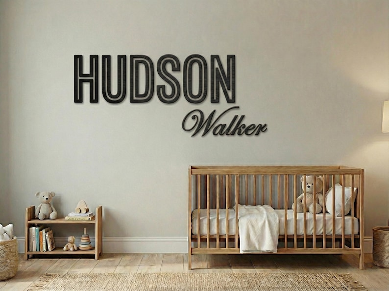 Puede incluir: Decoraci&oacute;n de pared de madera negra con las palabras "HUDSON" y "Walker" en un cuarto de ni&ntilde;os. Tambi&eacute;n se ve una cuna de madera con un oso de peluche y una peque&ntilde;a estanter&iacute;a con juguetes y libros.