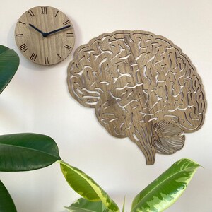 Puede incluir: Un reloj de pared de madera con n&uacute;meros romanos y un dise&ntilde;o de corte en forma de cerebro. El reloj est&aacute; montado en una pared blanca con una planta verde en primer plano.