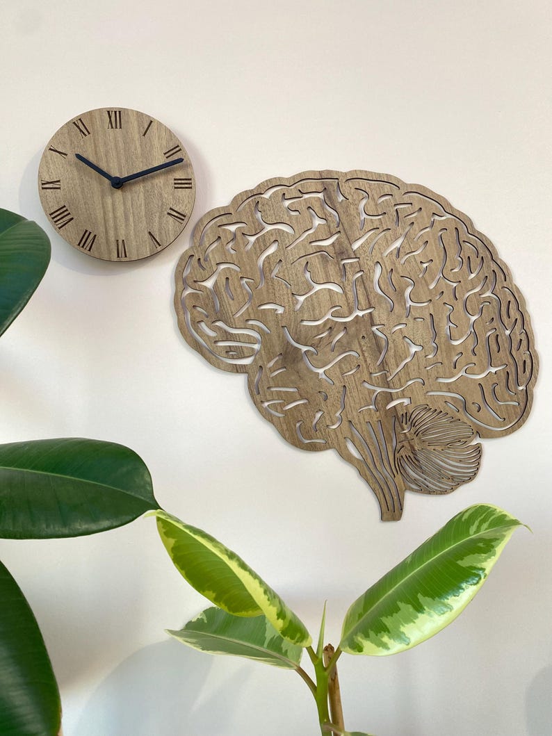 Puede incluir: Un reloj de pared de madera con n&uacute;meros romanos y un dise&ntilde;o de corte en forma de cerebro. El reloj est&aacute; montado en una pared blanca con una planta verde en primer plano.