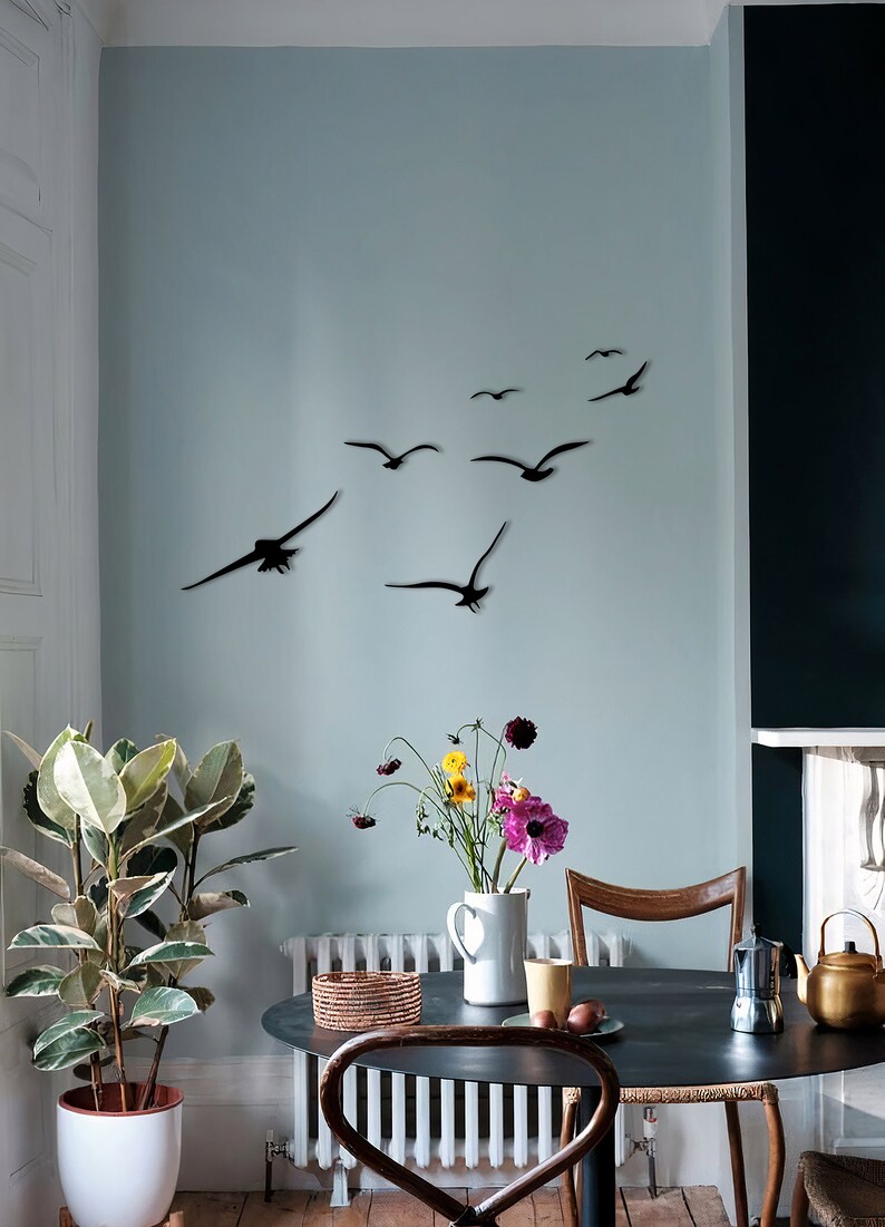 Puede incluir: Un comedor con una mesa y sillas negras. La pared est&aacute; pintada de un color azul claro y presenta una bandada de p&aacute;jaros de metal negro en vuelo. Una planta en maceta se encuentra en el suelo junto a la mesa.