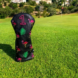 Puede incluir: Cubierta de cabeza de palo de golf negra con un patrón de flamencos rosas y el número "1" en letras rosas.