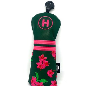 May include: A green golf club head cover with a pink floral design. The cover has a pink embroidered "H" in a circle and two pink stripes. A black tag is attached to the side.
