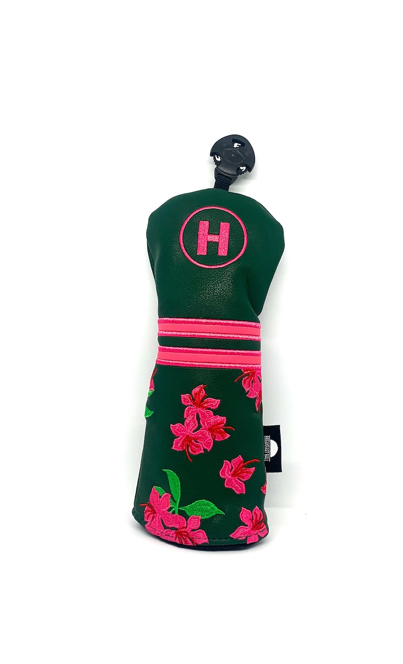 May include: A green golf club head cover with a pink floral design. The cover has a pink embroidered "H" in a circle and two pink stripes. A black tag is attached to the side.