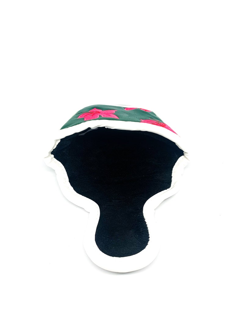May include: A dark green golf club head cover with embroidered pink floral designs. The cover has a white trim and a black interior. The cover is designed to protect golf clubs.