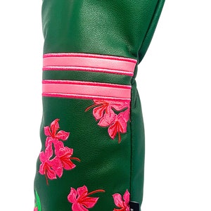 May include: Green golf club head cover with pink floral embroidery and a pink number 1 in a circle. The cover has pink stripes and a black tag with white text.