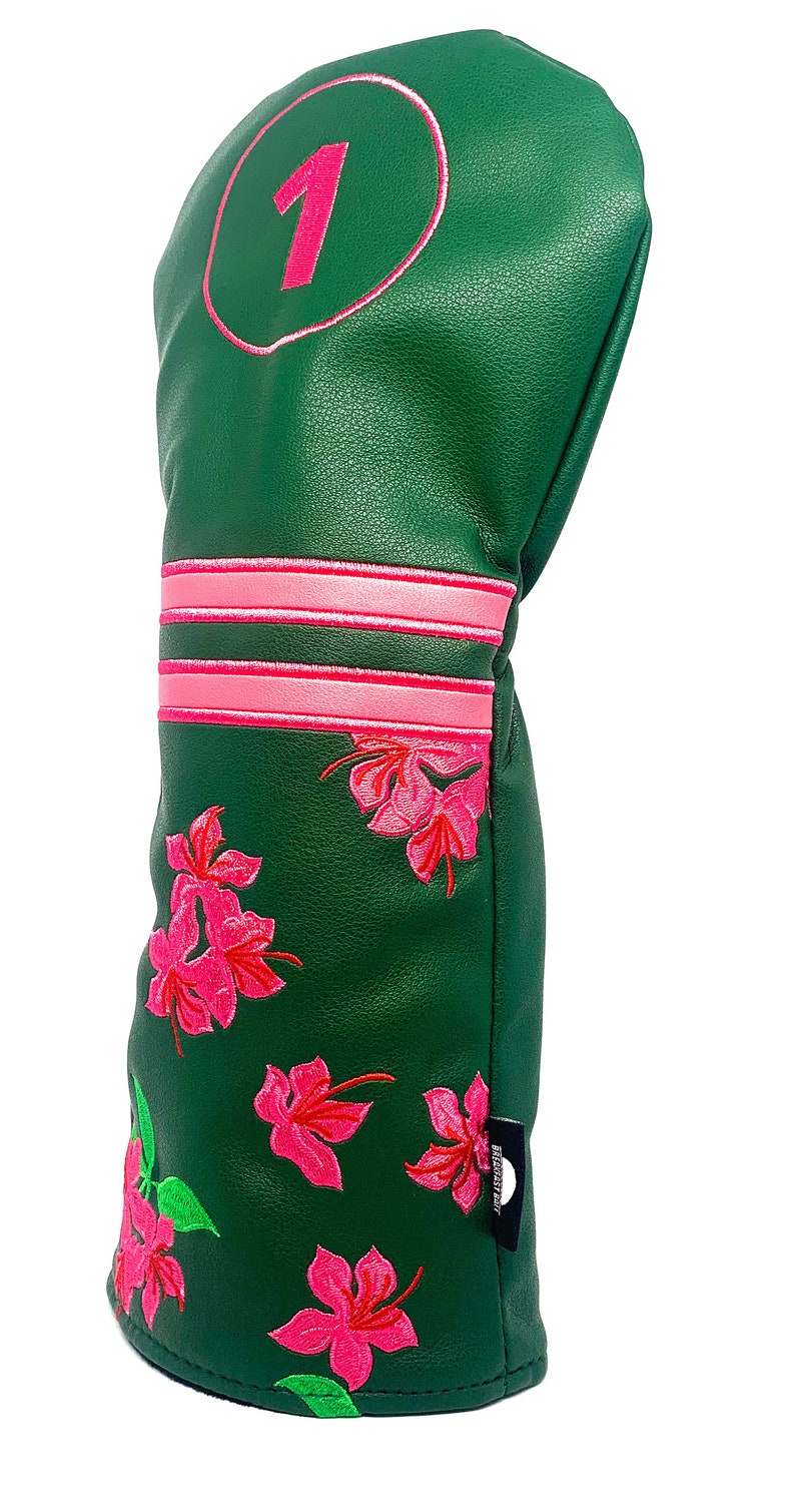 May include: Green golf club head cover with pink floral embroidery and a pink number 1 in a circle. The cover has pink stripes and a black tag with white text.