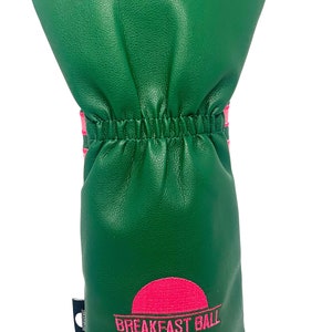 May include: Green golf club head cover with pink accents and the words "BREAKFAST BALL" in pink. The cover has a textured appearance and a gathered elastic band. The design includes a pink sun graphic.