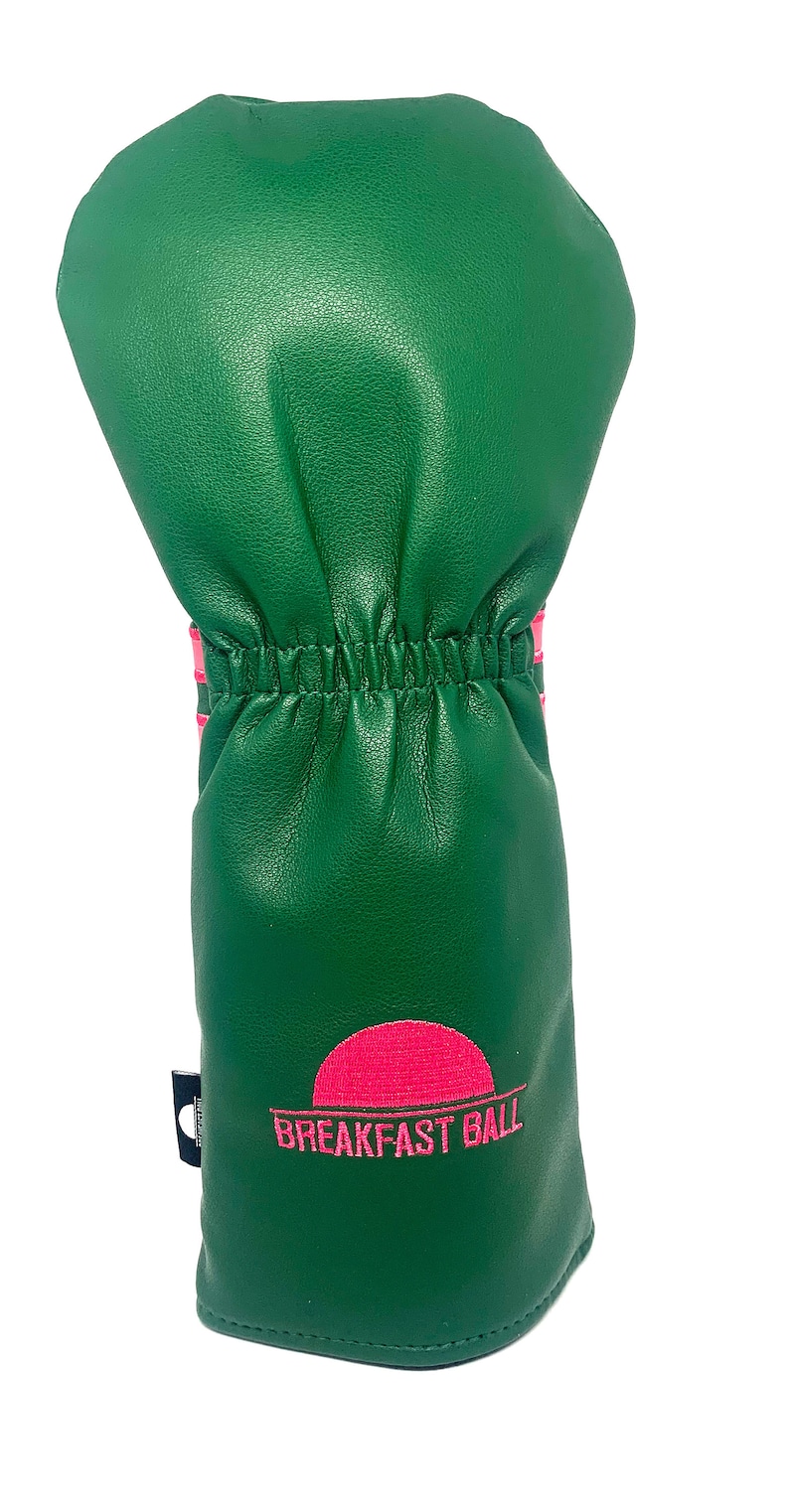 May include: Green golf club head cover with pink accents and the words "BREAKFAST BALL" in pink. The cover has a textured appearance and a gathered elastic band. The design includes a pink sun graphic.