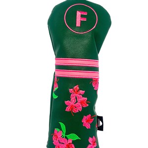 May include: A green golf club head cover with pink floral embroidery. The cover features a pink circle with the letter "F" and two pink stripes. A black clip is attached to the top.