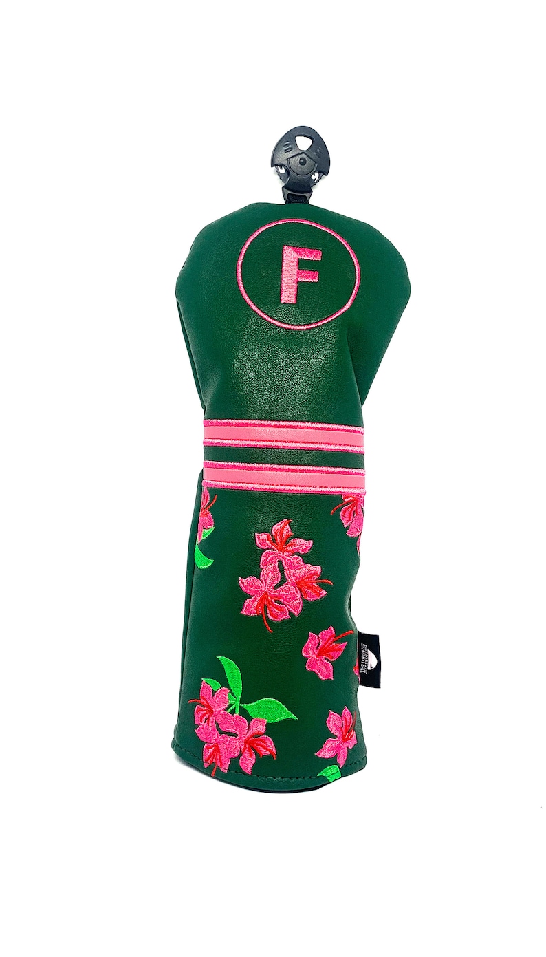 May include: A green golf club head cover with pink floral embroidery. The cover features a pink circle with the letter "F" and two pink stripes. A black clip is attached to the top.