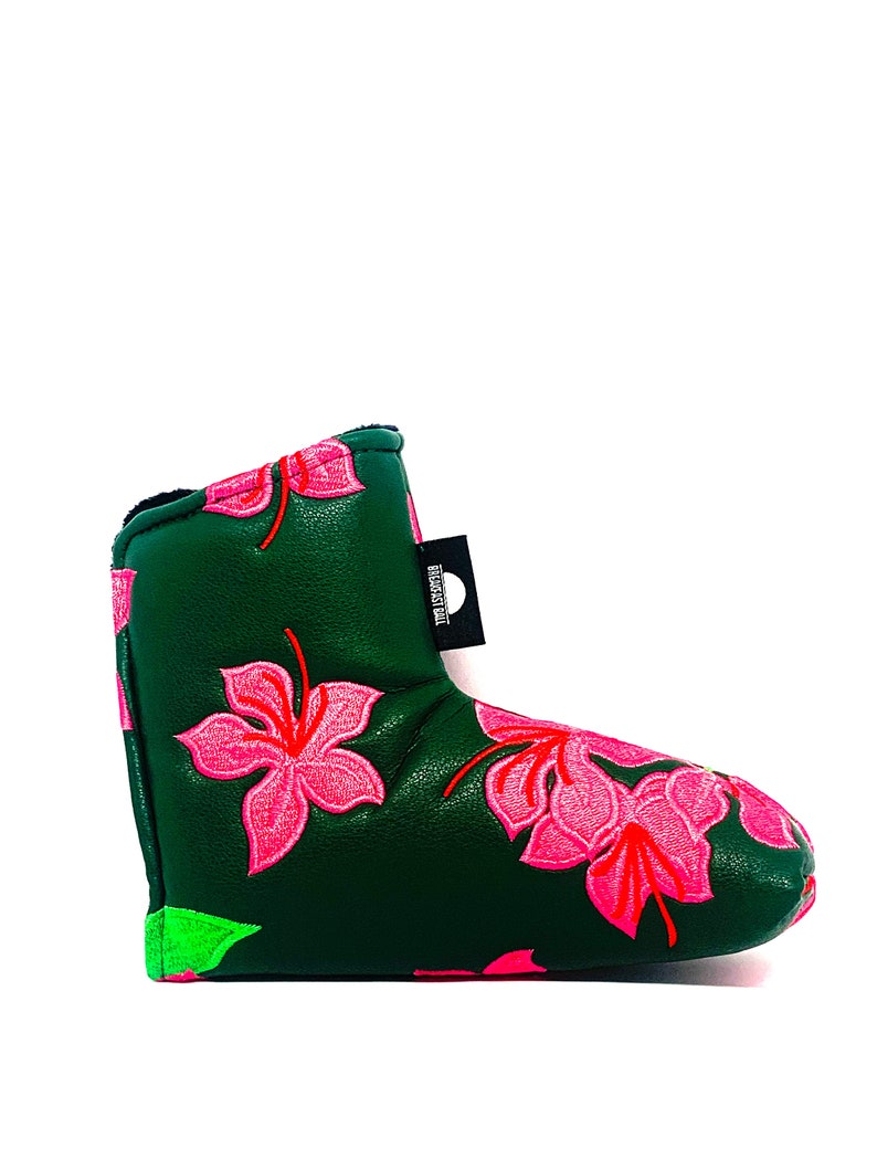 May include: A dark green golf club head cover with embroidered pink hibiscus flowers and red accents. The cover has a black tag with white text. The bottom edge is trimmed with neon green.