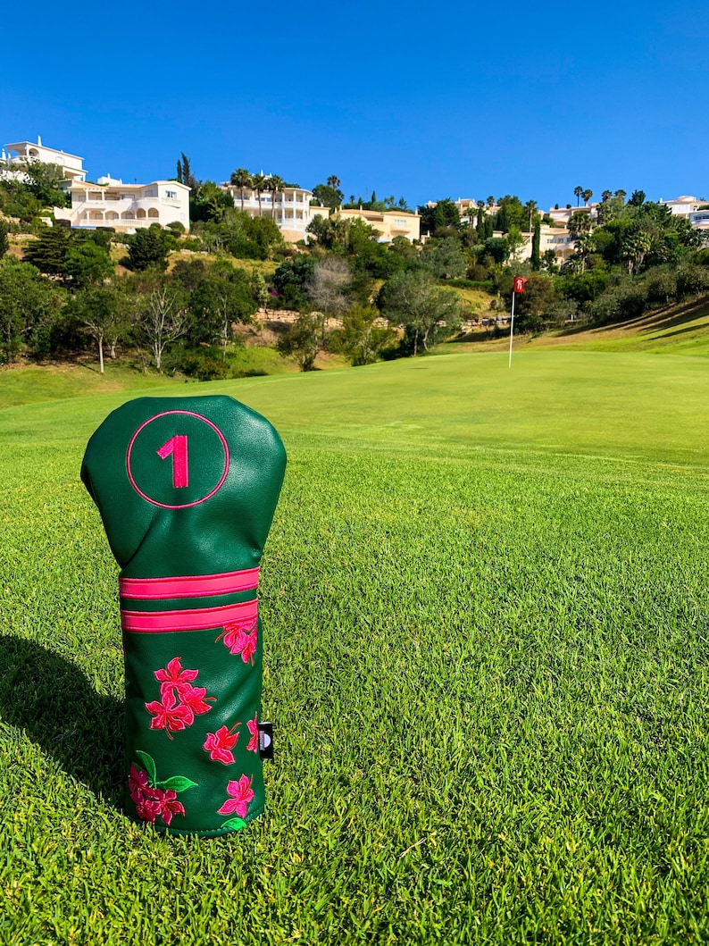 May include: A green golf club head cover with a pink number 1 and floral design. The cover is on a golf course with green grass and a blue sky. The cover has a pink trim and a small logo.