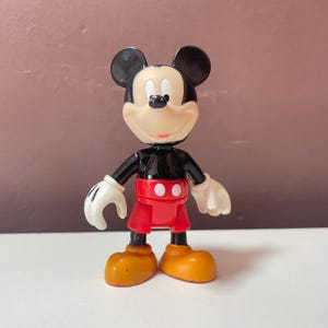May include: A plastic Mickey Mouse figurine featuring a black head with large round ears, a cream-coloured face, a black torso, red shorts with two white buttons, white gloves, and large yellow shoes. The character stands in an upright position.