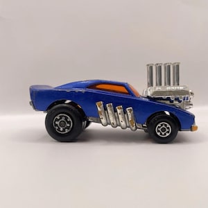 May include: A blue die-cast toy car featuring a prominent silver engine block protruding from the bonnet with four vertical exhaust pipes. The vehicle has orange-tinted windows, black wheels with silver rims, and silver side exhaust pipes.