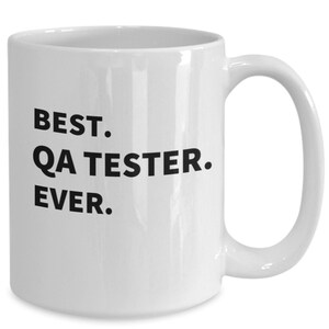 May include: A white ceramic mug with a smooth finish and a curved handle. The front of the mug features bold, black, sans-serif text printed in three lines that reads: BEST. QA TESTER. EVER. The design is centered on the exterior surface of the mug.