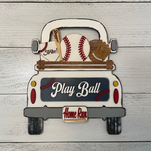May include: White wooden truck with a blue and red sign that says "Play Ball". The truck bed is filled with baseball equipment, including a baseball, a bat, a glove, and a baseball with the word "Strike!" on it. The truck has a license plate that says "Home Run".