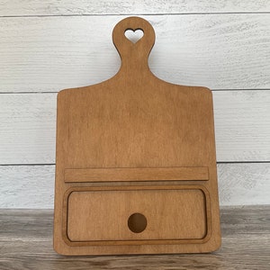 May include: A wooden cutting board with a heart-shaped cutout in the handle and a small drawer with a hole in the front. The board is stained a light brown color.