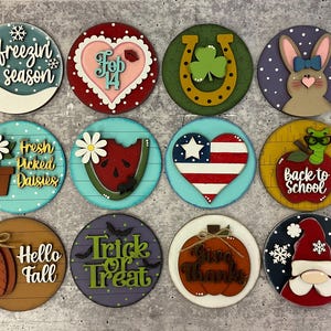 May include: A collection of twelve round wooden signs with various seasonal and holiday-themed designs. The signs feature images and text such as a snowflake, a heart, a horseshoe, a bunny, a daisy, a watermelon, an American flag heart, an apple, a pumpkin, and a gnome.