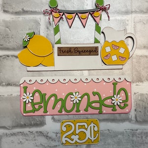 May include: A wooden lemonade stand sign with a pink and white lemonade sign, a yellow price tag that says "25 cents", and a yellow lemon graphic. The stand has a green and white striped roof with a banner that says "Fresh Squeezed".