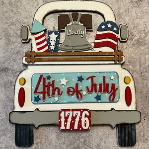 May include: White wooden truck with a red, white, and blue American flag, a red, white, and blue firework, a Liberty Bell, and a sign that says "4th of July" and "1776".
