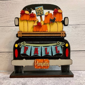 May include: A wooden fall-themed decoration featuring a black truck bed filled with orange pumpkins and hay bales. The truck has a banner that says "FALL" and a sign that says "Pick 25¢". The word "Pumpkins" is also displayed.