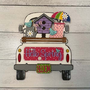 May include: A white wooden truck with a pink and white "Hello Spring" sign on the back. The truck is decorated with a birdhouse, a butterfly, two birds, a watering can, a rainbow umbrella, and rain boots. The license plate says "Bloom".