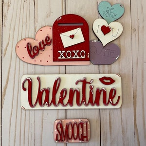 May include: A set of Valentine's Day decorations featuring a red mailbox with a white envelope, a pink heart with the word "love" in red, a purple heart with the word "cutie", a green heart with the words "be mine", a red heart with the words "I love you", a white sign with the word "Valentine" in red, and a pink sign with the word "smooch" in red.