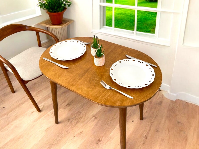 May include: A small, oval wooden dining table with a light brown finish. Two white plates with heart-shaped cutouts, silverware, and small potted plants are arranged on the table. A wooden chair is visible to the left.