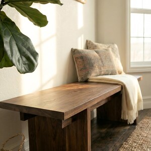 May include: A dark brown wooden bench with a rectangular top and a sturdy base. Two decorative pillows and a cream-colored throw blanket rest on the bench. The bench is placed near a window and a large green plant.