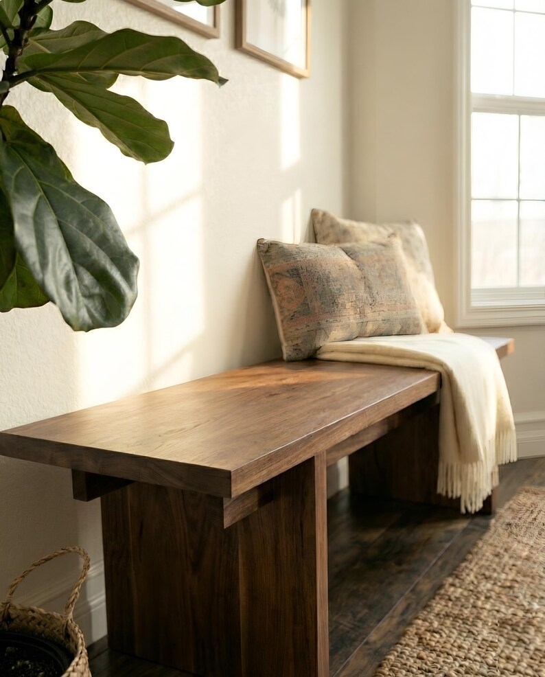 May include: A dark brown wooden bench with a rectangular top and a sturdy base. Two decorative pillows and a cream-colored throw blanket rest on the bench. The bench is placed near a window and a large green plant.