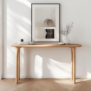 May include: A light-colored wooden console table with a curved top and four rounded legs. A framed abstract art print hangs above the table, which is decorated with a small sculpture, books, and a vase with branches.