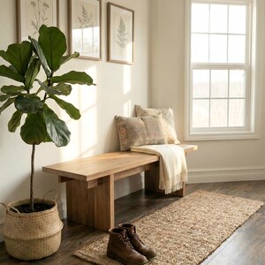 May include: A light wood bench with two decorative pillows and a cream-colored throw blanket. A woven jute rug and brown leather boots sit on the dark wood floor. A potted plant and framed botanical prints complete the interior decor.