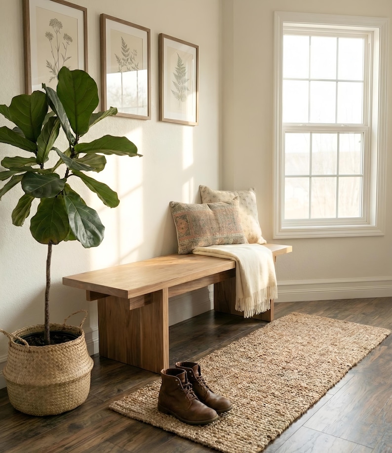 May include: A light wood bench with two decorative pillows and a cream-colored throw blanket. A woven jute rug and brown leather boots sit on the dark wood floor. A potted plant and framed botanical prints complete the interior decor.