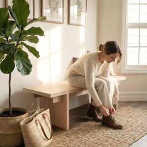 May include: A light wood bench with a person tying their brown leather boots. A woven jute rug and a large potted plant in a basket are in the foreground. Framed botanical prints hang on the wall.