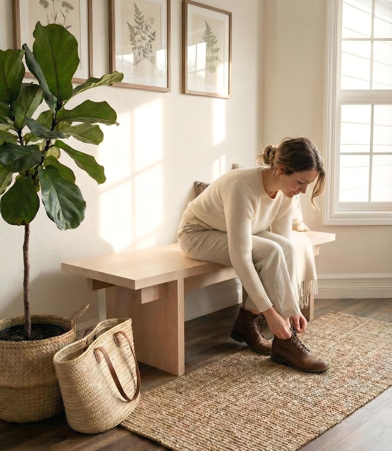 May include: A light wood bench with a person tying their brown leather boots. A woven jute rug and a large potted plant in a basket are in the foreground. Framed botanical prints hang on the wall.