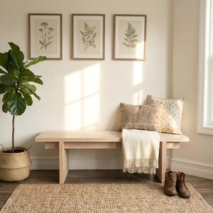 May include: A cozy interior scene featuring a light wood bench with pillows and a fringed throw. Botanical prints hang above. A woven rug and a potted plant complete the inviting space. Brown leather boots sit on the floor.