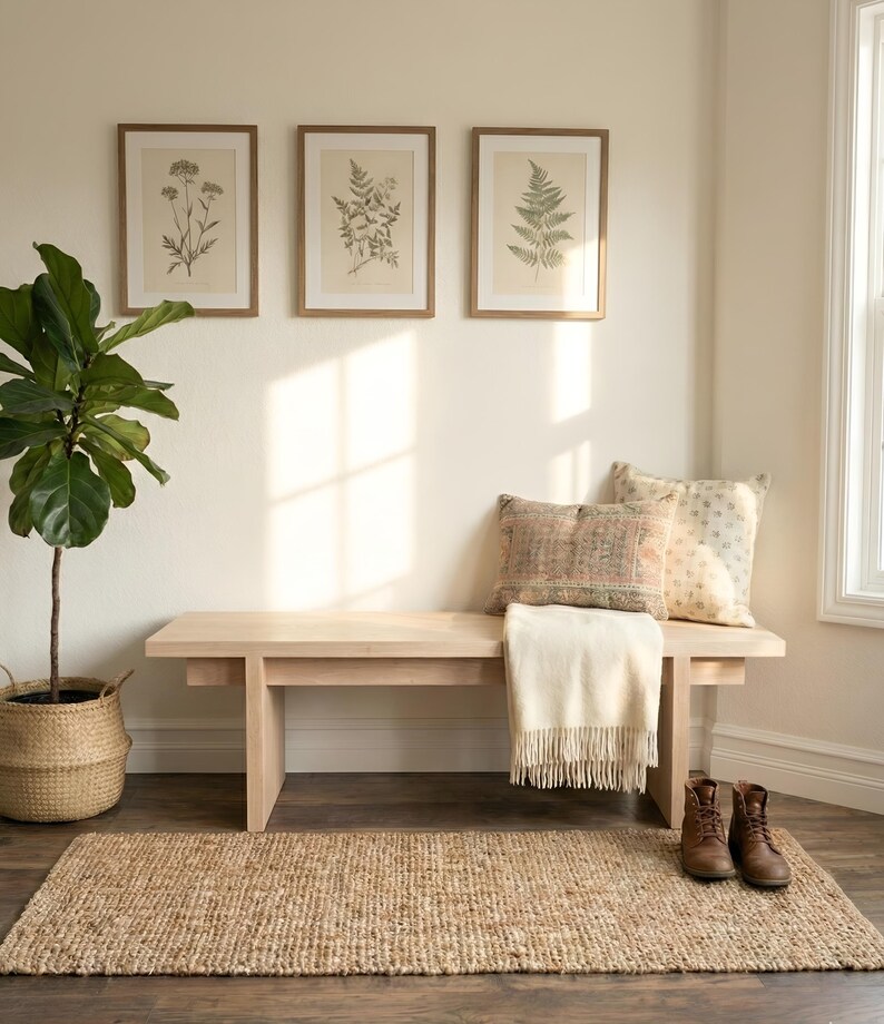 May include: A cozy interior scene featuring a light wood bench with pillows and a fringed throw. Botanical prints hang above. A woven rug and a potted plant complete the inviting space. Brown leather boots sit on the floor.